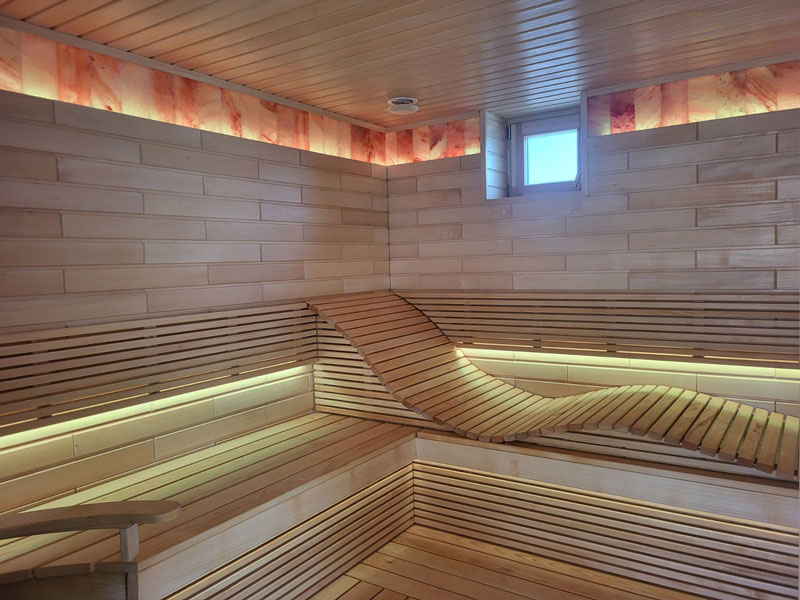 Světlá dřevěná sauna s vlnitou lavicí a LED osvětlením.