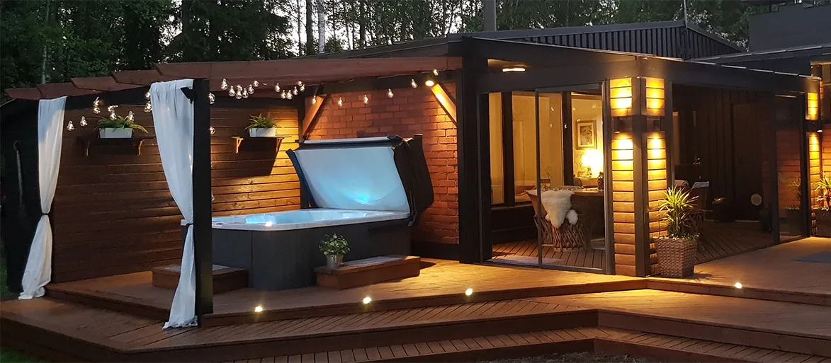 An evening terrace with lighting and a gazebo, equipped with an outdoor jacuzzi, next to a cozy cottage with glass doors