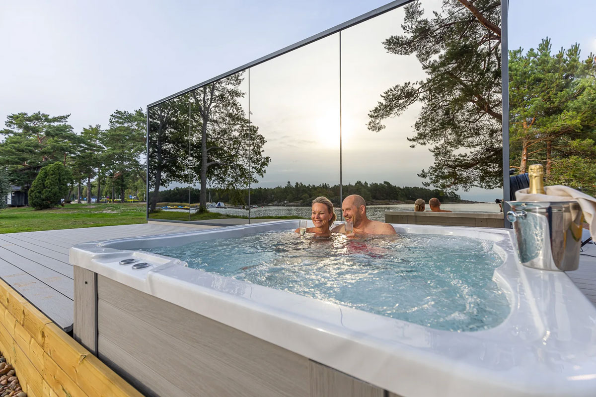 A couple enjoys an outdoor jacuzzi by a mirrored wall, holding champagne glasses in their hands, with the sunset by the lake providing the backdrop